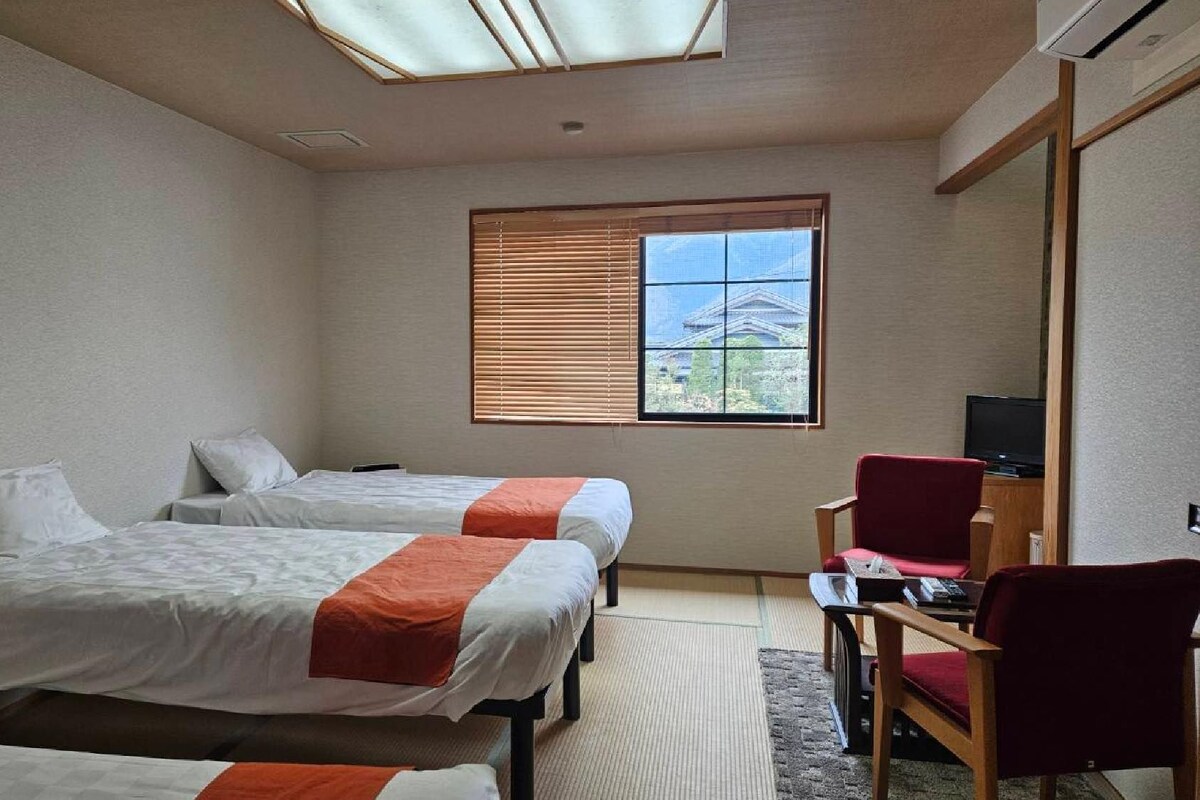 A hotel room with two beds, a television, a red chair, and a window with blinds.