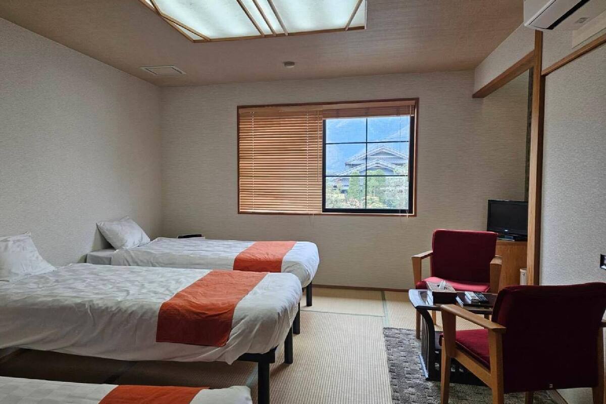 A hotel room with two beds, a television, a red chair, and a window with blinds.