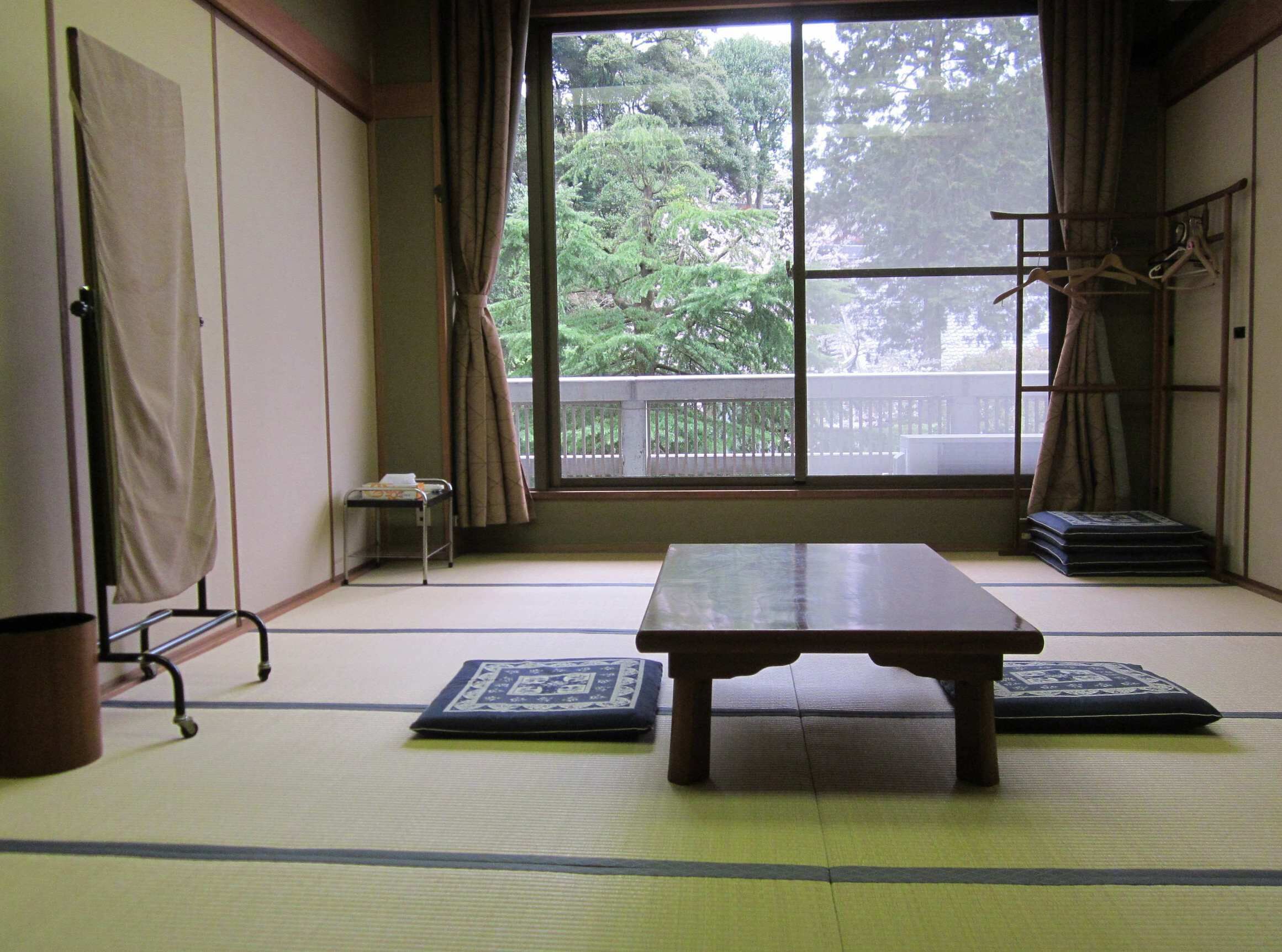 Habitació japonesa tradicional amb sòl de tatami, taula de fusta baixa i vista a un balcó amb vegetació.