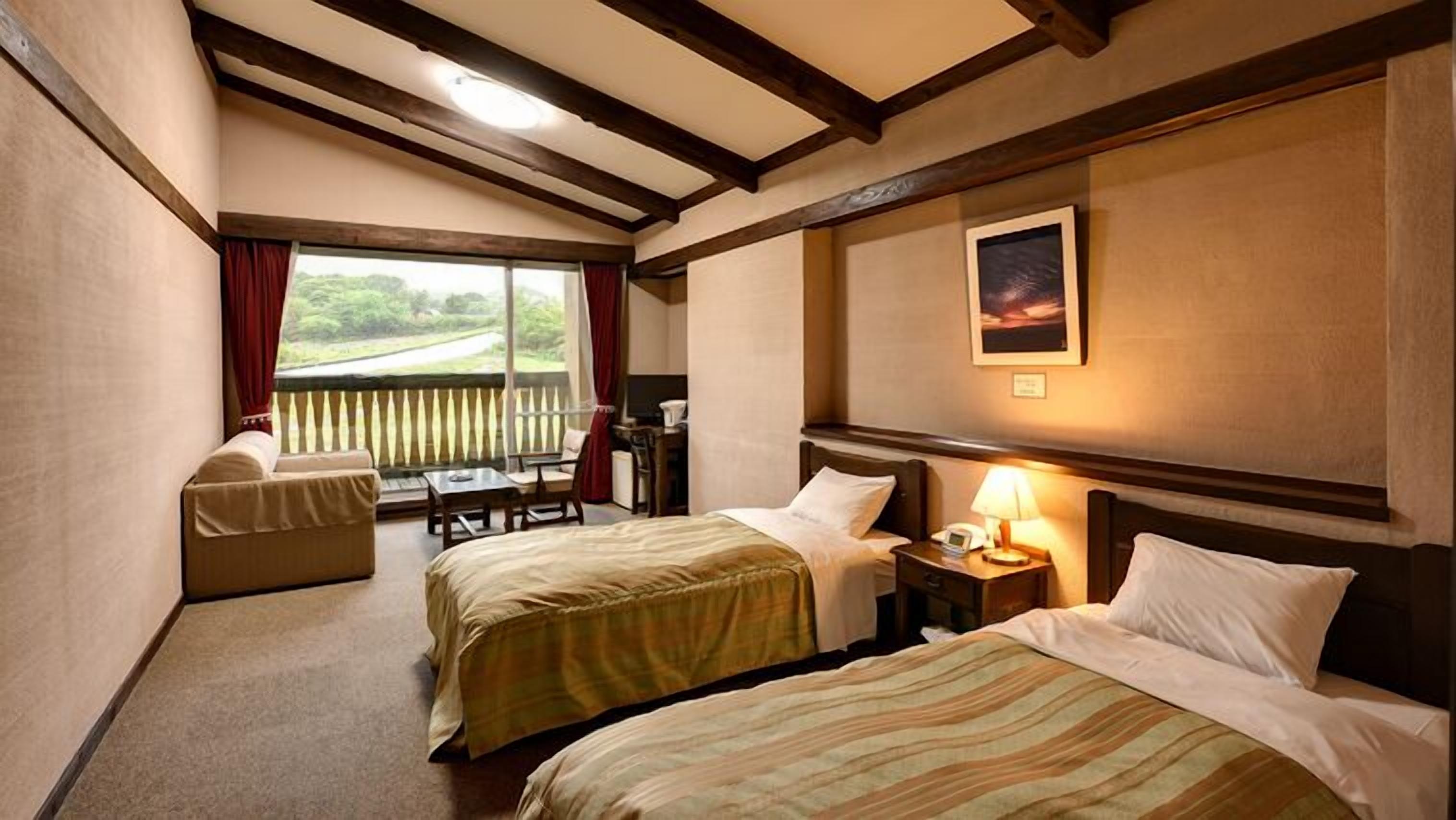 A hotel room with two beds, a small table, a chair, a window with a view, and a painting on the wall.