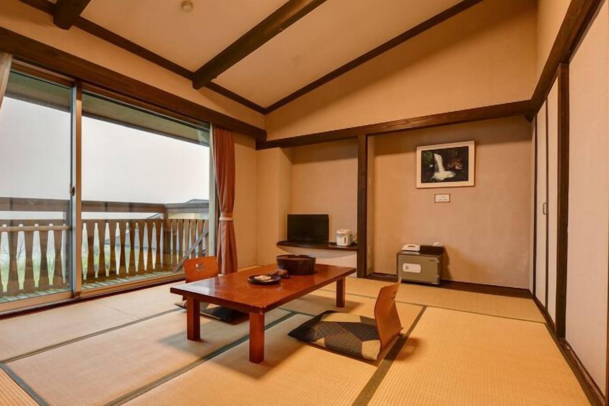 Ein traditioneller japanischer Raum mit Tatami-Boden, einer Schiebetür zu einem Balkon, einem Fernseher und einem gerahmten Bild an der Wand.