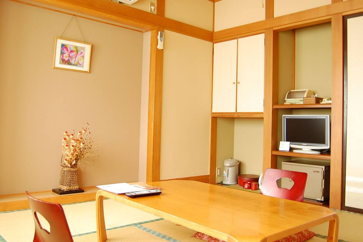 A traditional Japanese-style room with tatami flooring, a wooden table, a red chair, a television, and a small cabinet.