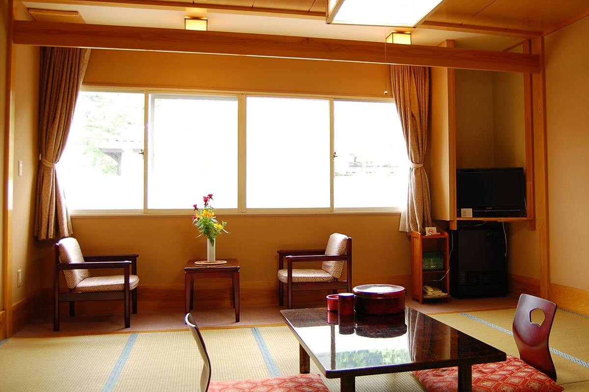 A room with tatami flooring, a low table, two chairs, a TV, and a window with curtains.