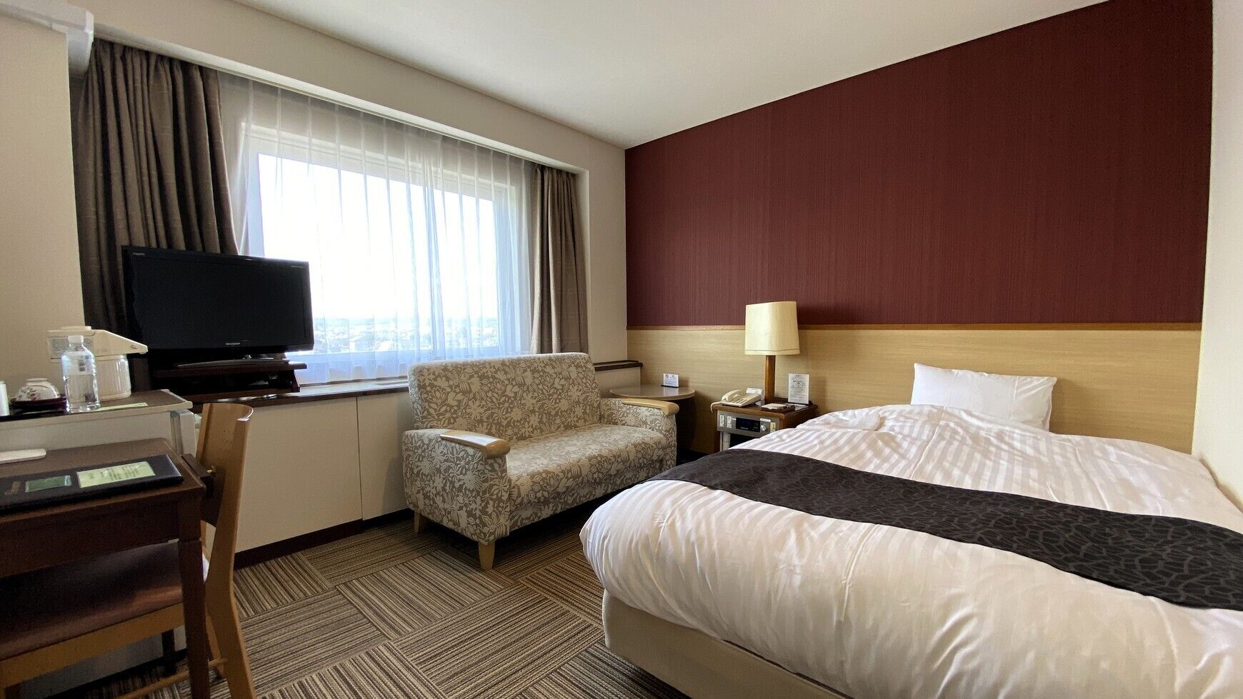 A hotel room with a bed, a desk, a chair, a television, and a window with curtains.