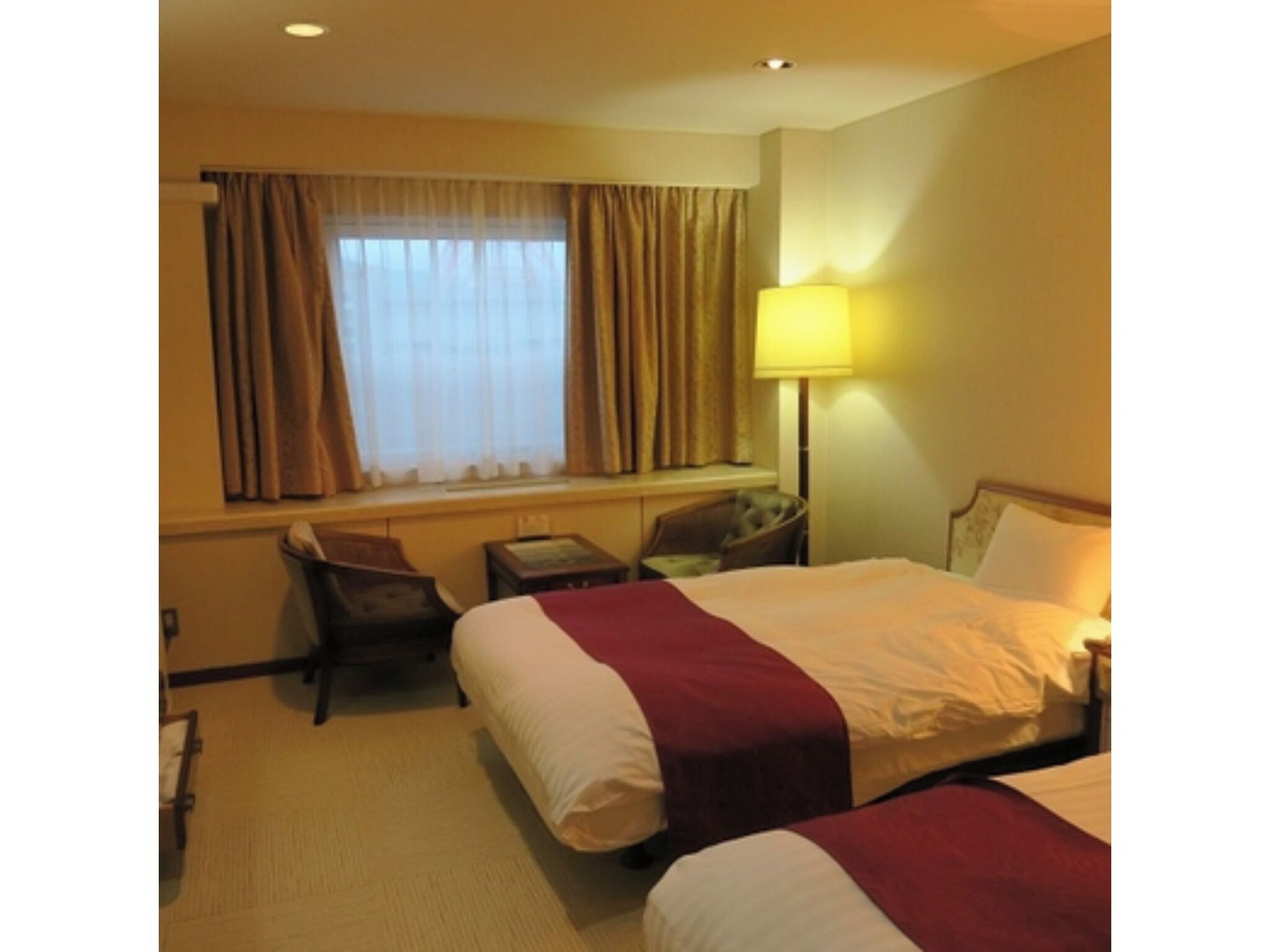 A hotel room with two beds, a desk, a chair, a lamp, and a window with curtains.