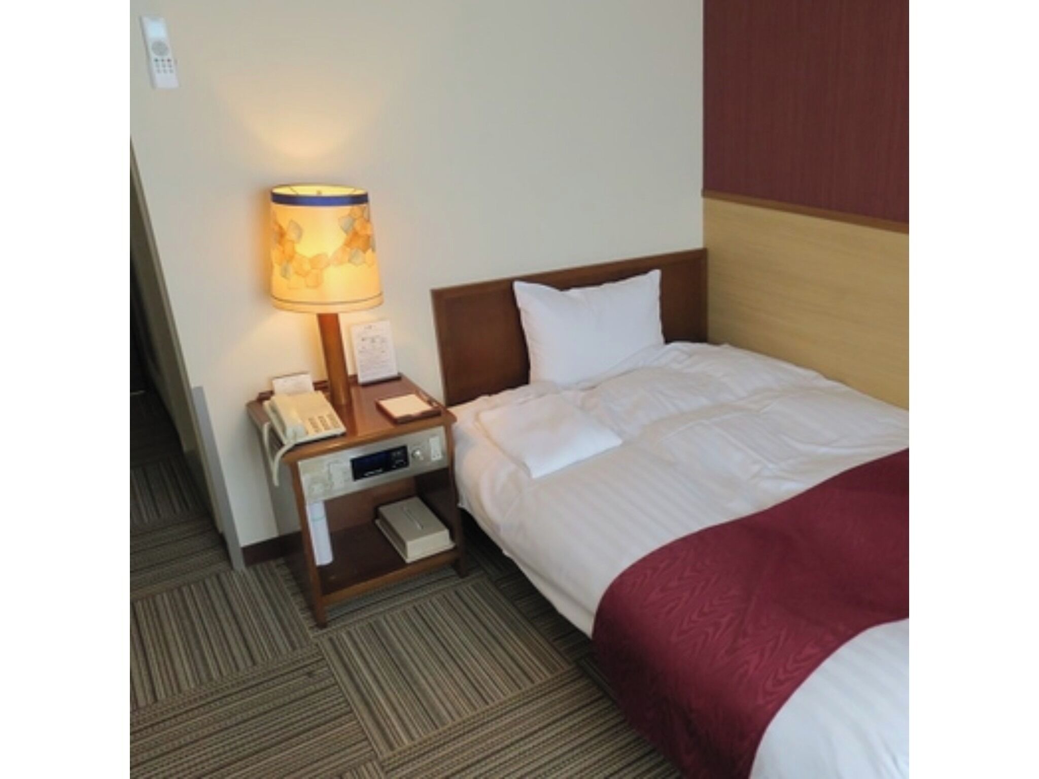 A hotel room with a neatly made bed, a bedside lamp, a telephone, and a small table with a book and a notepad.