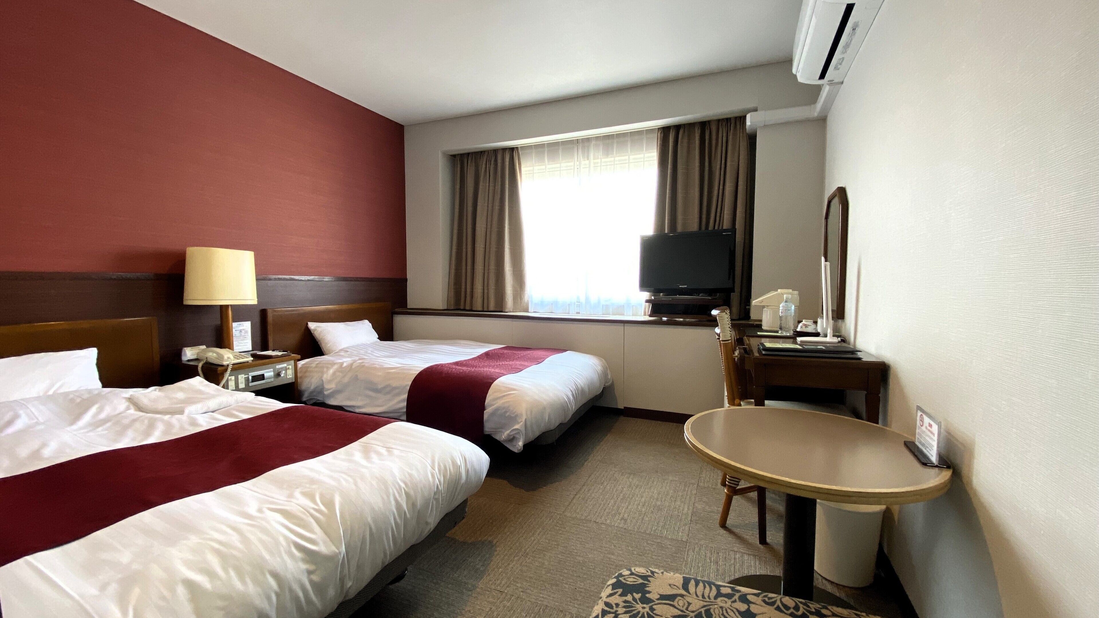 A hotel room with two beds, a desk, a television, a mirror, and an air conditioning unit.