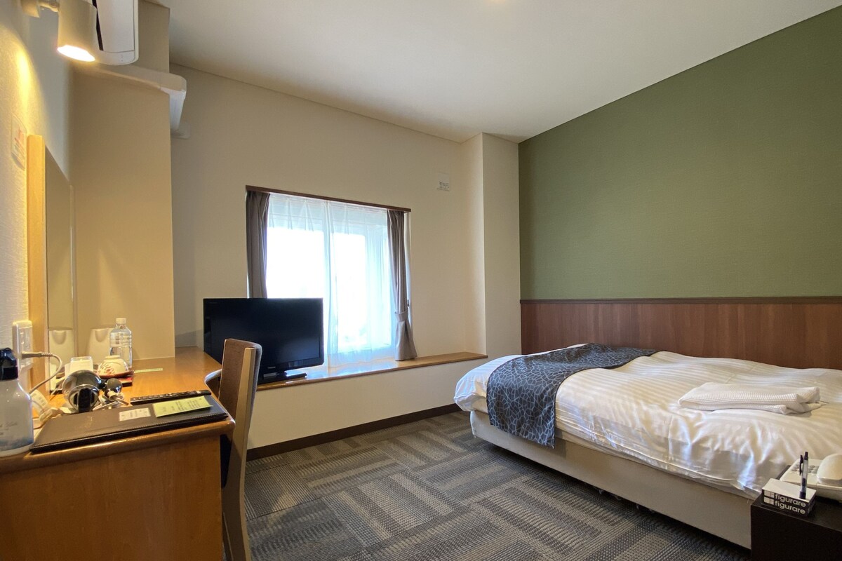 Habitación de hotel con cama, escritorio con ordenador, silla, televisor y ventana con cortinas.