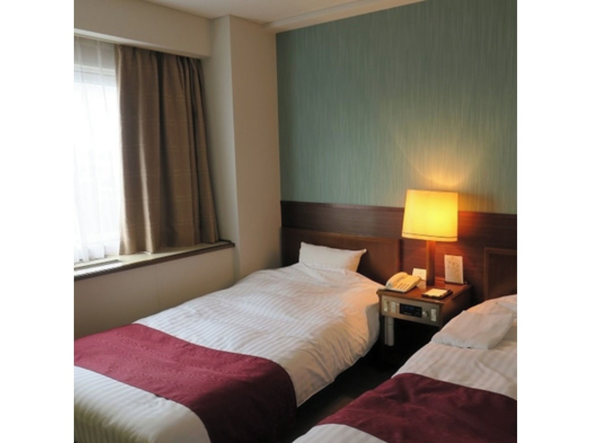 A hotel room with two beds, a nightstand with a lamp, and a window with curtains.