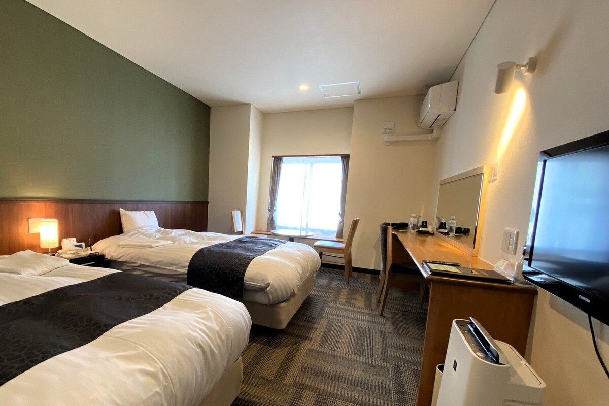 Habitación de hotel con dos camas, un escritorio con televisor, una silla y una ventana.