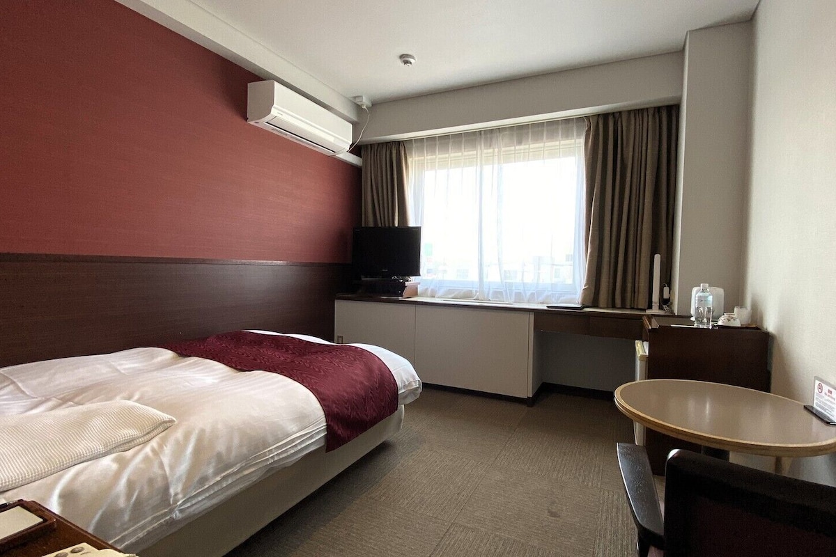 Habitación de hotel con cama, escritorio, silla, mesita pequeña, unidad de aire acondicionado, televisor y ventana con cortinas.
