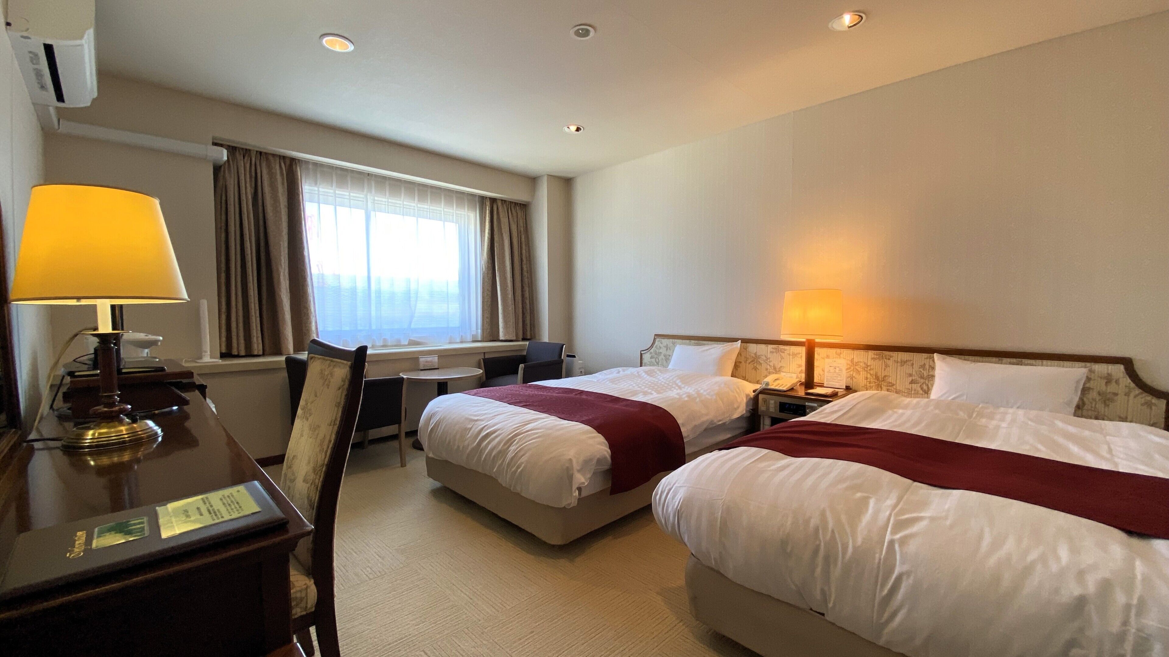 A hotel room with two beds, a desk, a chair, a lamp, a window with curtains, and a wall-mounted air conditioner.