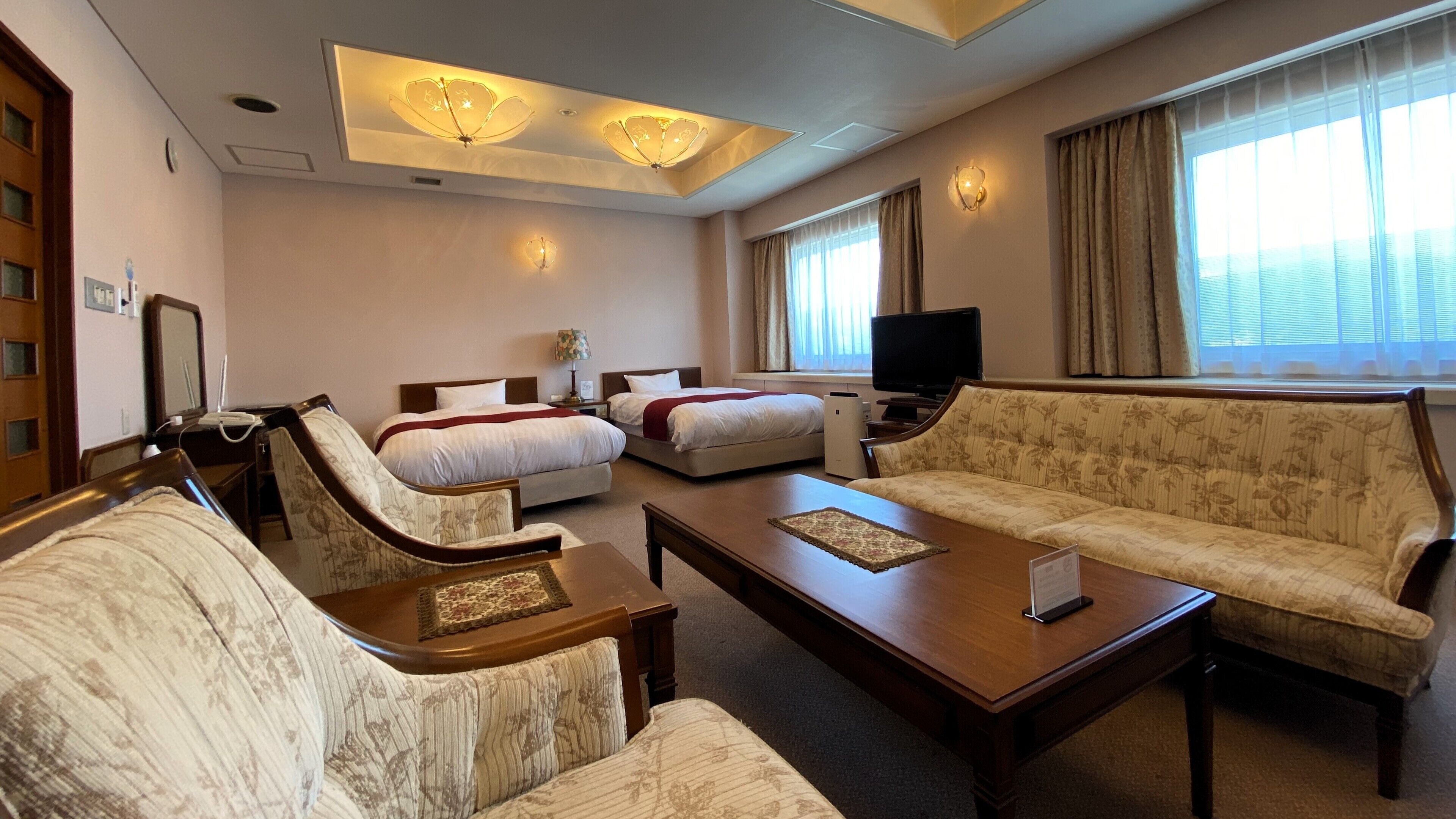 A hotel room with a bed, two armchairs, a sofa, a coffee table, a TV, and a large window with curtains.