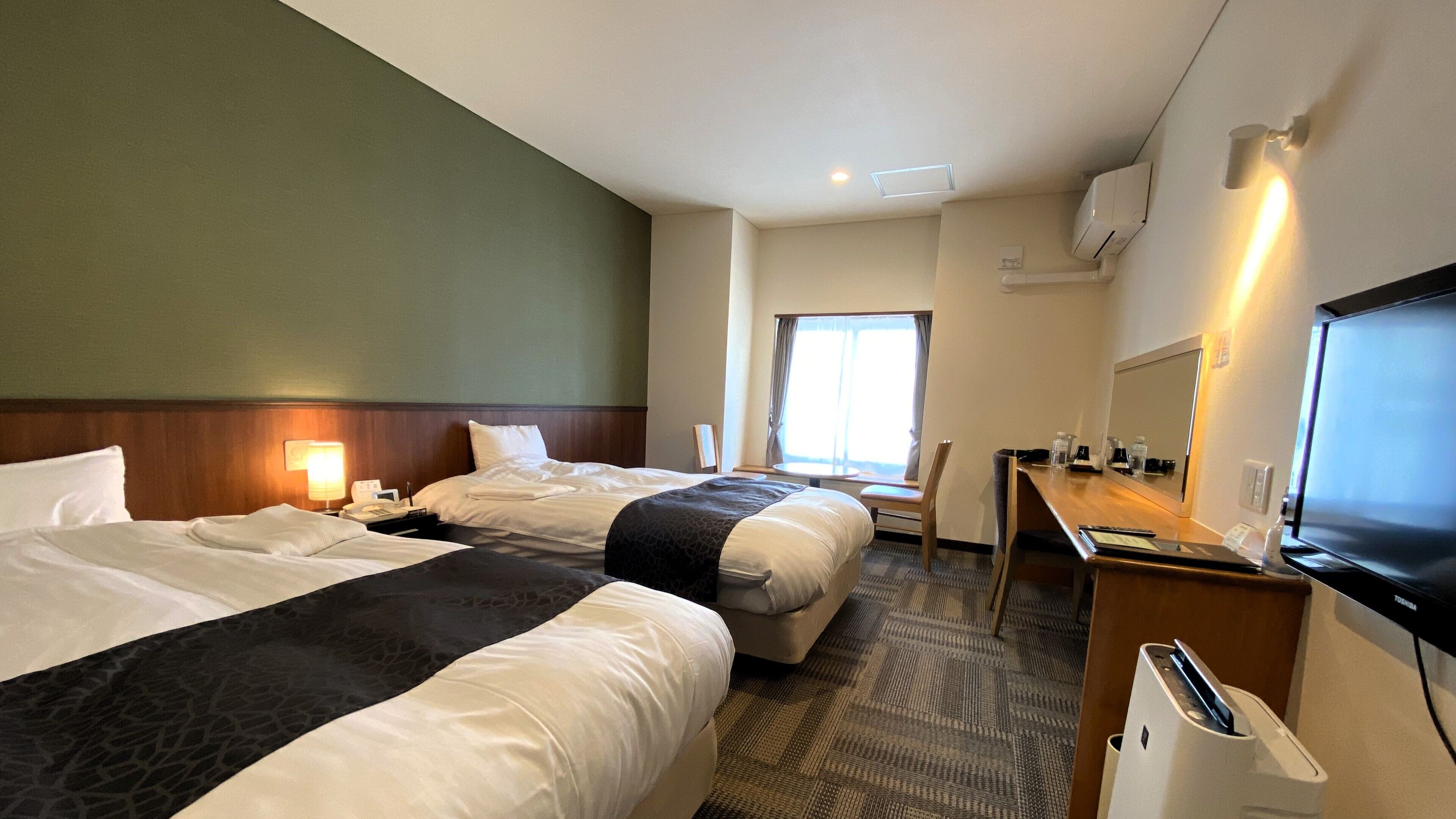 A hotel room with two beds, a desk, a television, and a window.