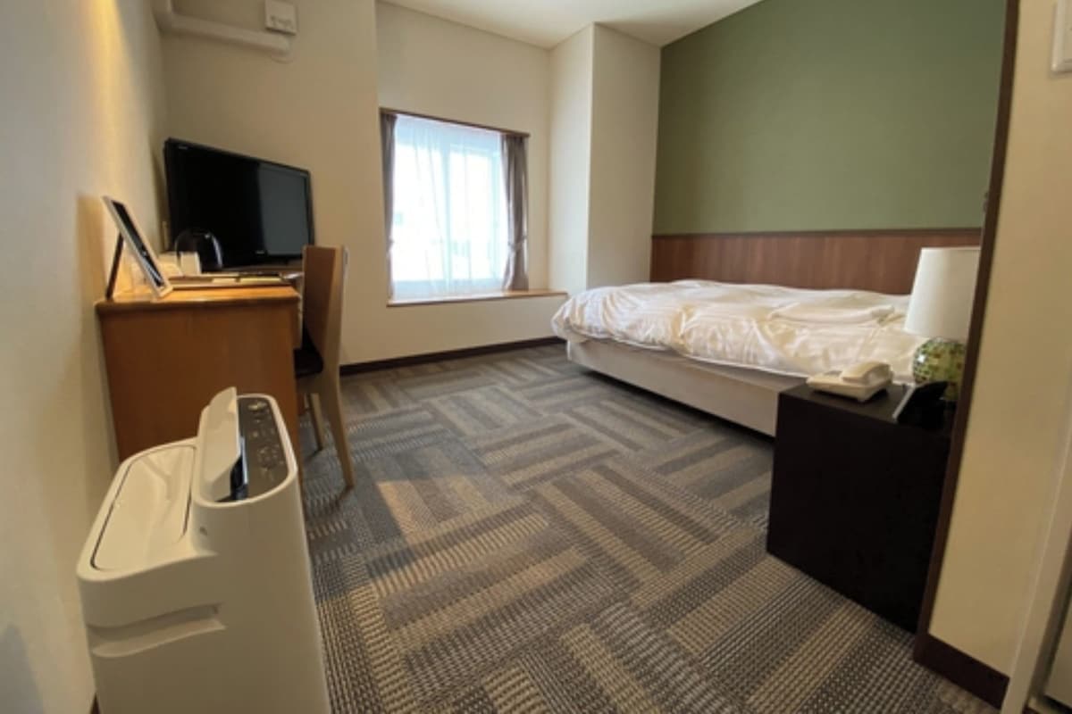 Una habitación de hotel con una cama, un escritorio con computadora, una silla, un cesto de basura y una ventana con cortinas.