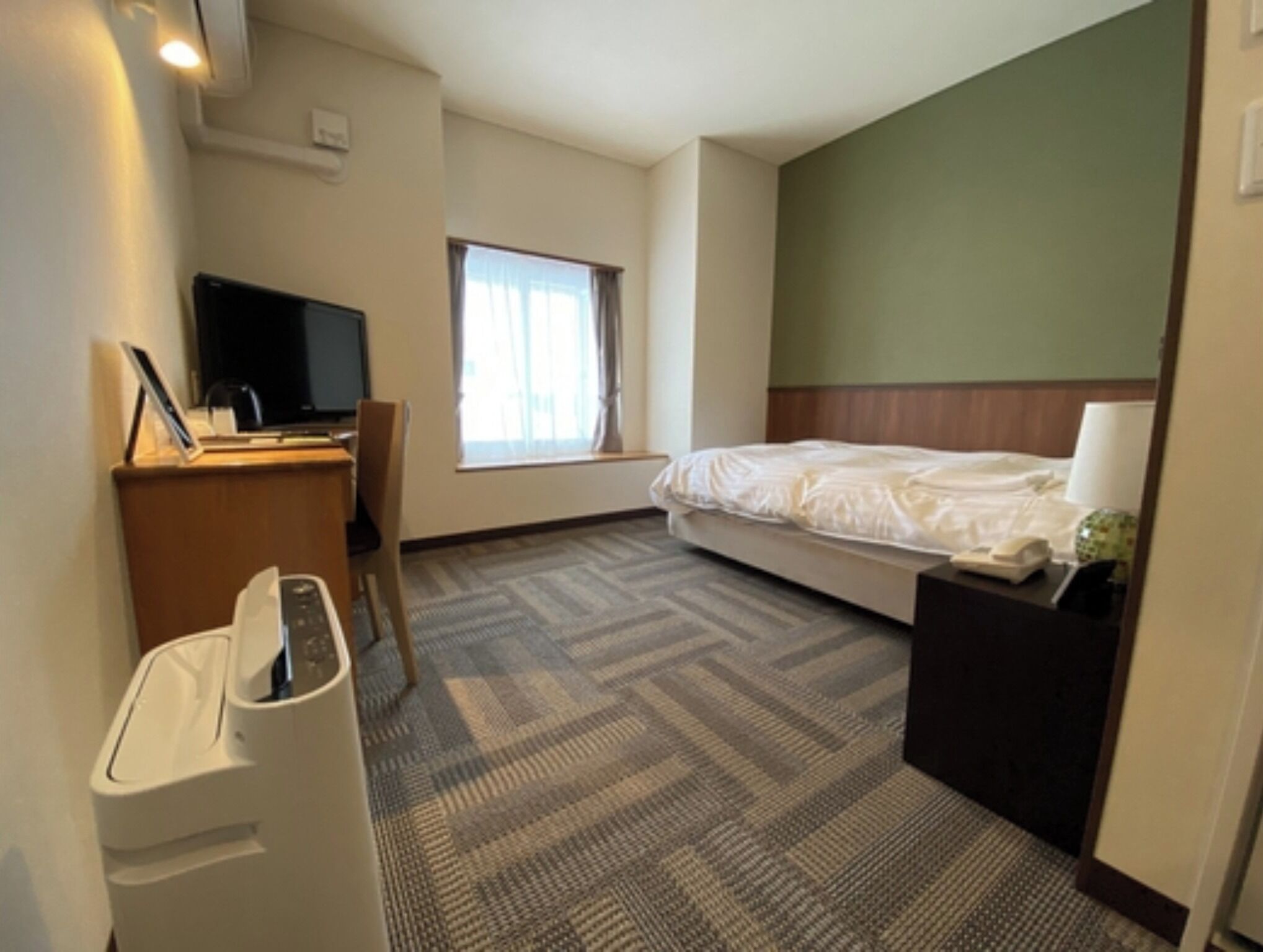 A hotel room with a bed, a desk with a computer, a chair, a waste bin, and a window with curtains.
