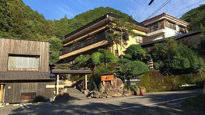 Il s’agit d’un bâtiment japonais traditionnel, avec une façade en bois, un chemin de pierre menant à l’entrée et une petite structure ressemblant à un sanctuaire située à l’avant.