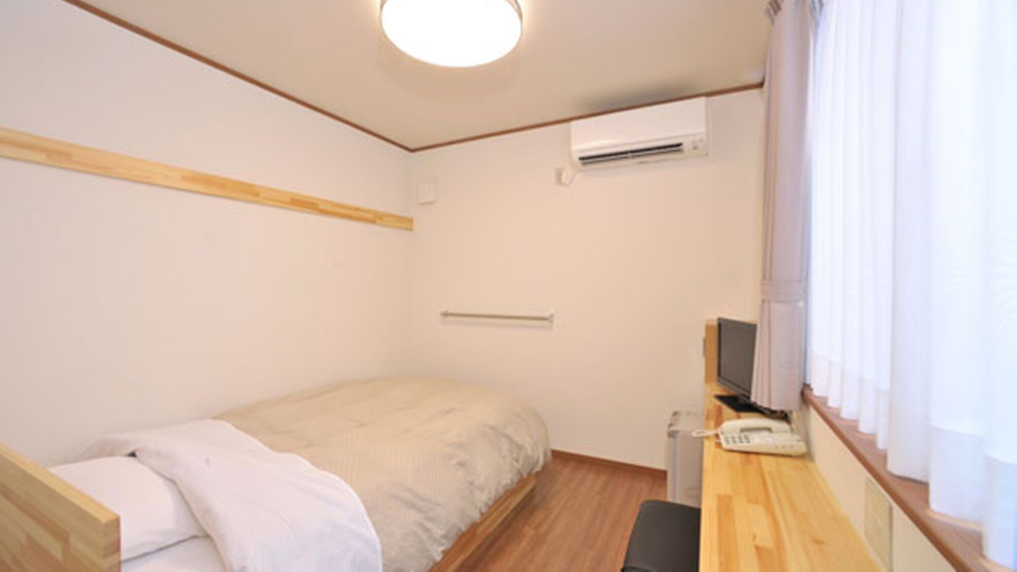 Una habitación pequeña con una cama individual, cabecera de madera, escritorio con teléfono y aire acondicionado en la pared.