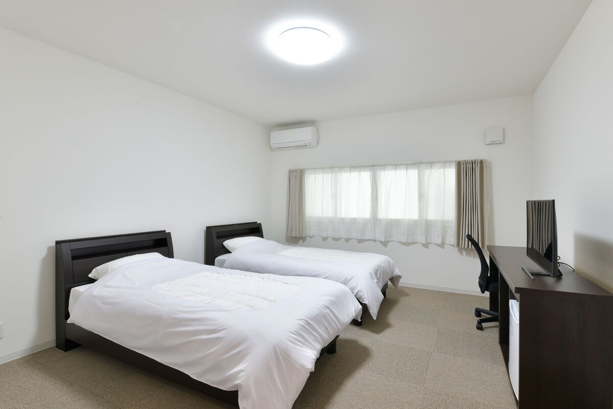 A hotel room with two beds, a desk, a television, and a window with curtains.