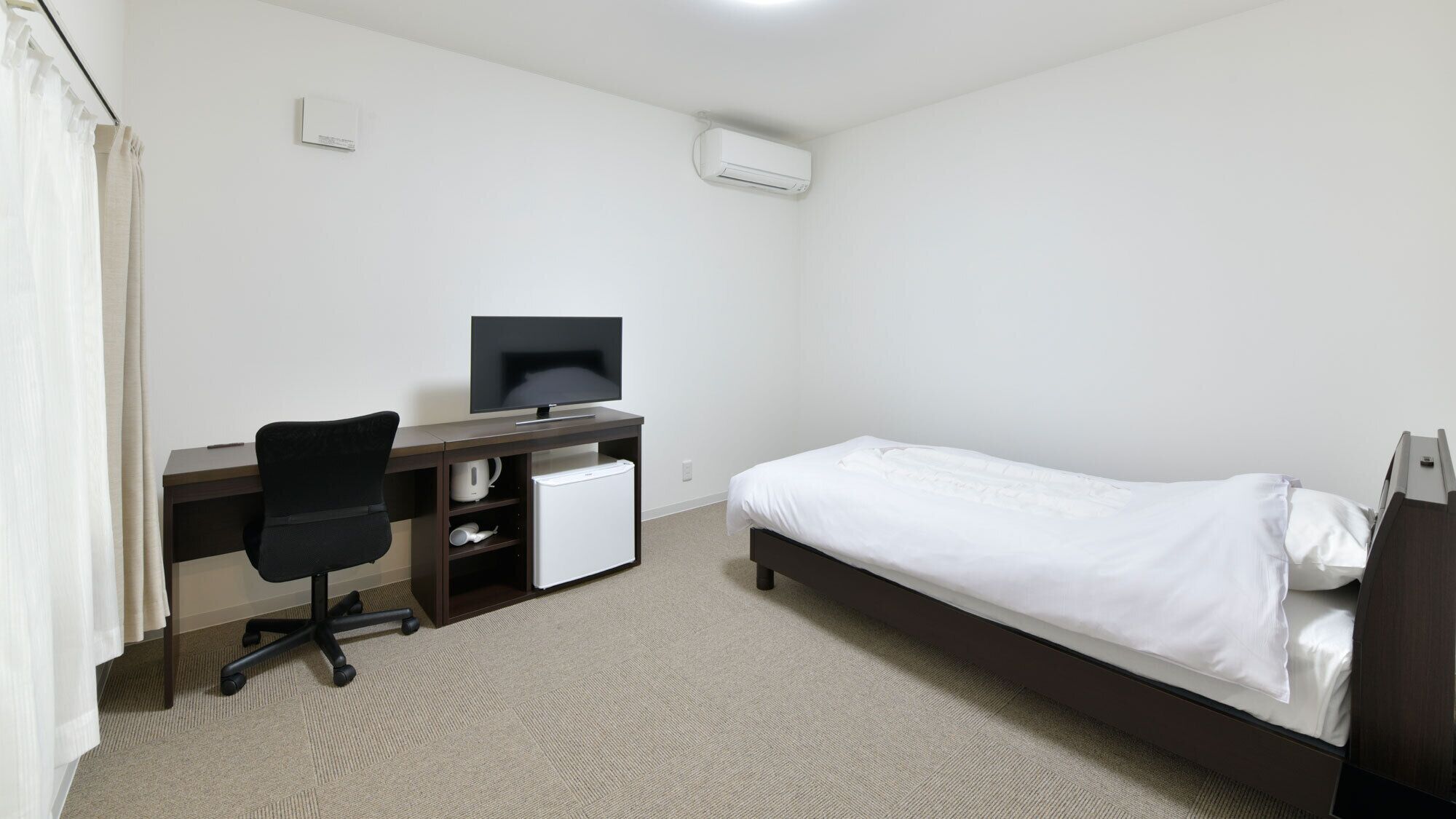 Habitación de hotel con cama, escritorio con computadora, silla, refrigerador pequeño y unidad de aire acondicionado.