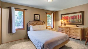 Une chambre à coucher avec un lit, deux fenêtres avec des rideaux, une table de chevet avec une lampe, une commode et un tableau au mur.