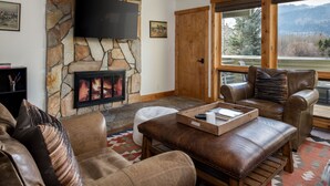 A living room with a stone fireplace, leather furniture, a flat-screen TV, and a large window with a mountain view. - Outpost: Windflower 2115 - Mountain Views! (Wilson)