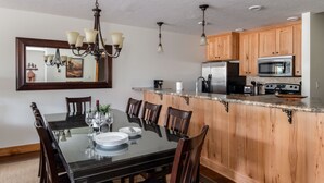 A dining area with a glass table and wooden chairs, a kitchen with wooden cabinets and stainless steel appliances, and a chandelier above the table. - Outpost: Windflower 2115 - Mountain Views! (Wilson)
