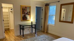 A bedroom with a bed, a desk with a lamp, a painting, a window with curtains, and a door. - Farmhouse Retreat (Rougemont)