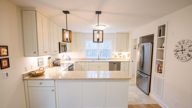 A modern kitchen with white cabinets, a central island, stainless steel appliances, pendant lighting, and a clock on the wall. - Farmhouse Retreat (Rougemont)