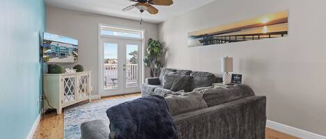 A living room with a grey sofa, a ceiling fan, a TV on the wall, and a large window with a view of buildings.