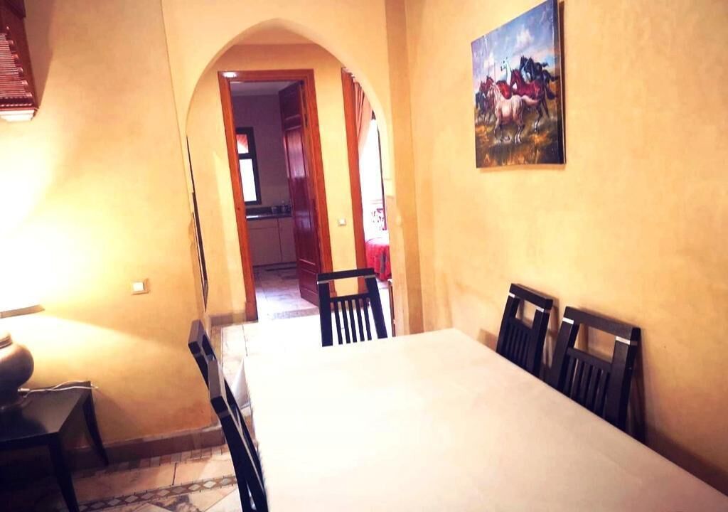 Un espace repas avec une longue table et des chaises, une arche donnant accès à une autre pièce, et un tableau accroché au mur.