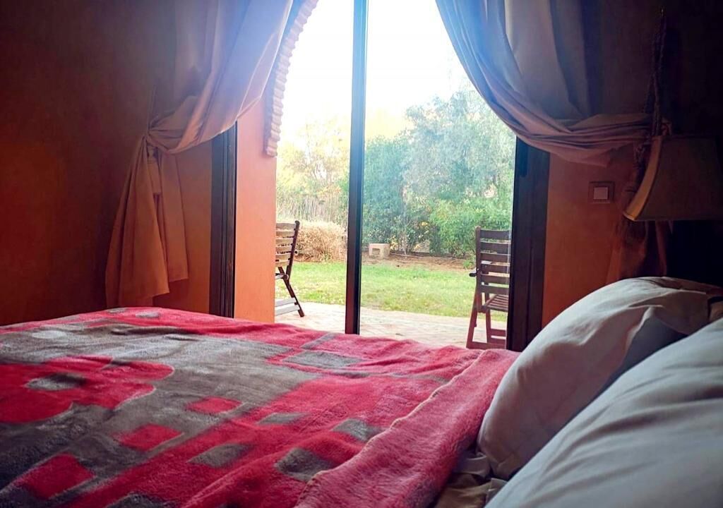 A bedroom with a bed, a red and gray blanket, and a view of an outdoor area with a chair and greenery.