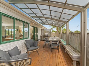 A covered patio with a dining table set for four, surrounded by wicker chairs and a wooden bench. - The Sunny Bach - Cooks Beach Holiday Home (Cooks Beach)