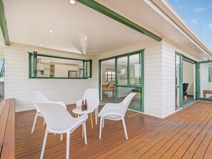 A covered outdoor area with wooden flooring, white wicker furniture, and a glass door leading to an interior space. - The Sunny Bach - Cooks Beach Holiday Home (Cooks Beach)