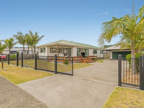 A single-story house with a covered porch, a front yard with a small garden, and a black metal fence. - The Sunny Bach - Cooks Beach Holiday Home (Cooks Beach)