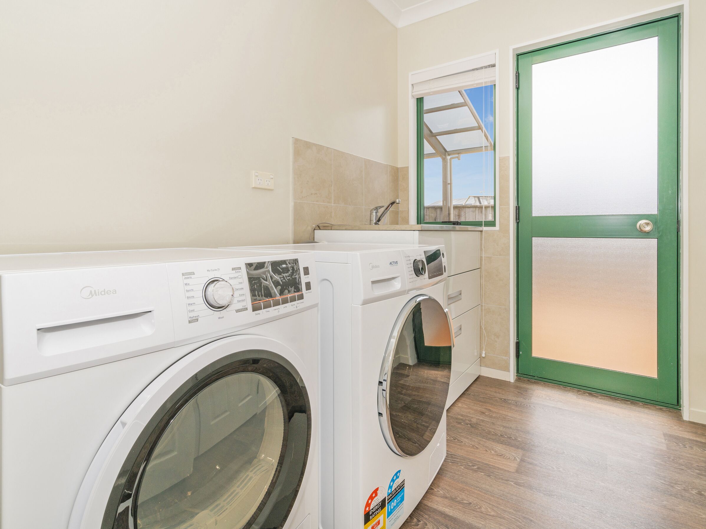 A laundry room with two Midea washing machines, a green door, and a window above the washing machines.