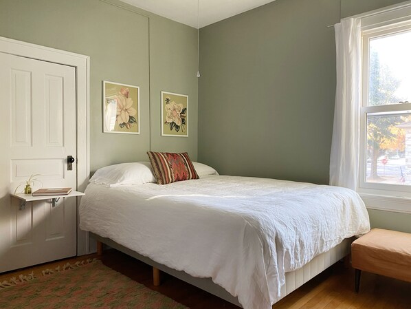Un dormitorio con una cama, una mesita de noche con un libro, una ventana con vistas y dos cuadros colgados en la pared.