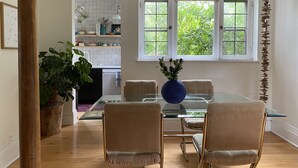 A dining area with a glass table, four chairs, and a vase of flowers. A kitchen is visible in the background with a window showing greenery outside. - Eclectic Home Near UWM Campus and Lake Michigan (Shorewood)