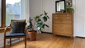 A room with a wooden floor, a chair with a patterned cushion, a wooden dresser with drawers, a potted plant, and a window with curtains. - Eclectic Home Near UWM Campus and Lake Michigan (Shorewood)