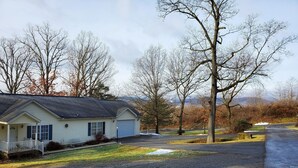 A single-story house with a garage, surrounded by trees and a gravel driveway. - Cottage at Chimney Ridge Farms (Duncansville)