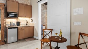 Una cucina compatta con microonde, macchina del caffè e lavandino. C'è un tavolo da pranzo in legno con due sedie e una bottiglia di vino. Una porta scorrevole conduce a un'altra stanza.