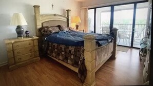 Une chambre à coucher comprenant un lit, des tables de chevet, une lampe, une croix accrochée au mur et un balcon aménagé avec du mobilier d’extérieur.