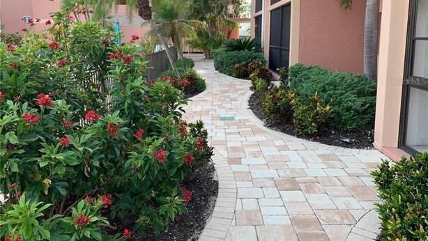Un chemin menant à un bâtiment dont l’entrée est ornée de carreaux, entouré d’une végétation luxuriante et de plantes en fleurs.