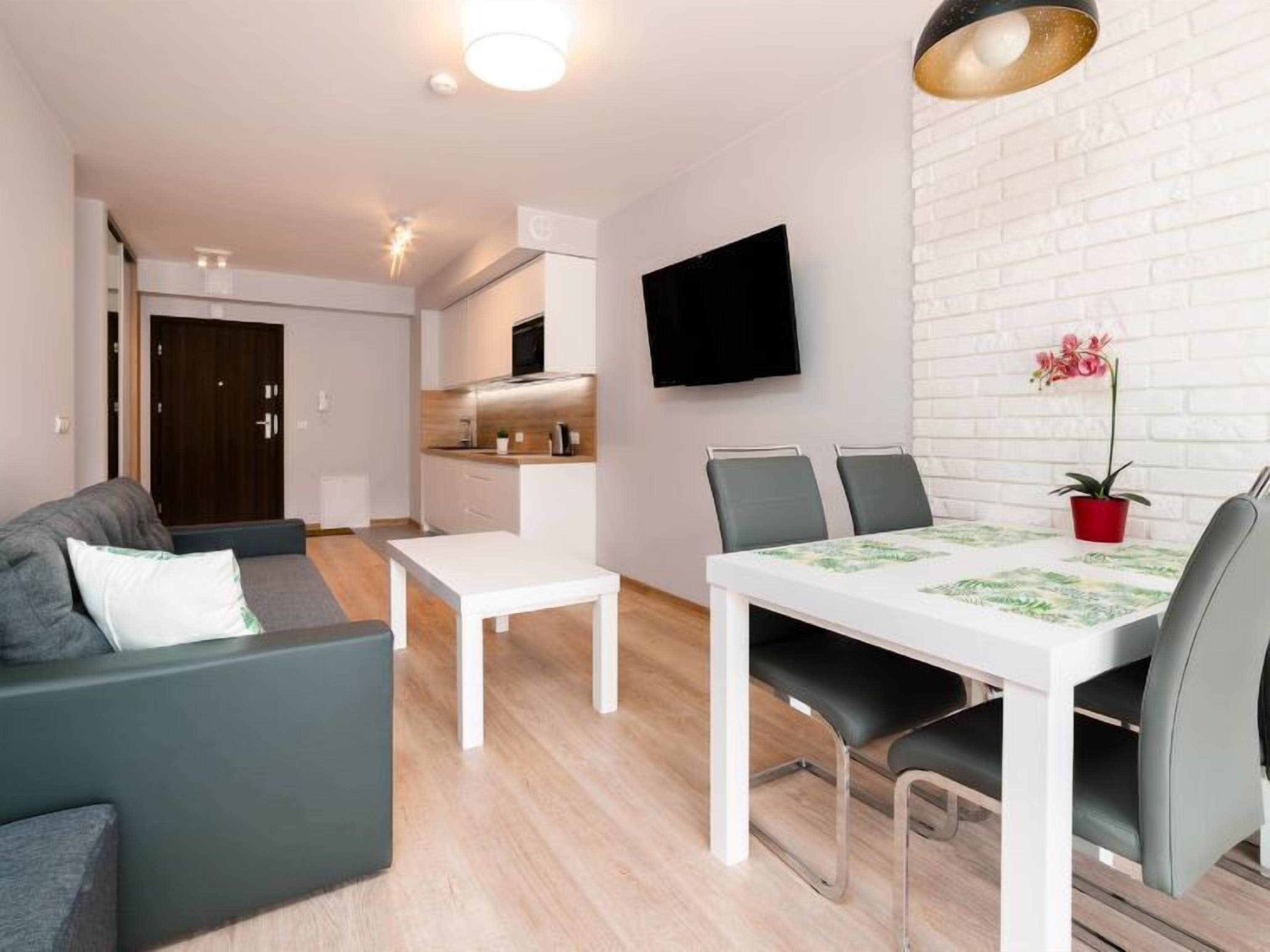 A modern living room with a sofa, a dining table with chairs, a flat-screen TV mounted on the wall, and a potted plant.