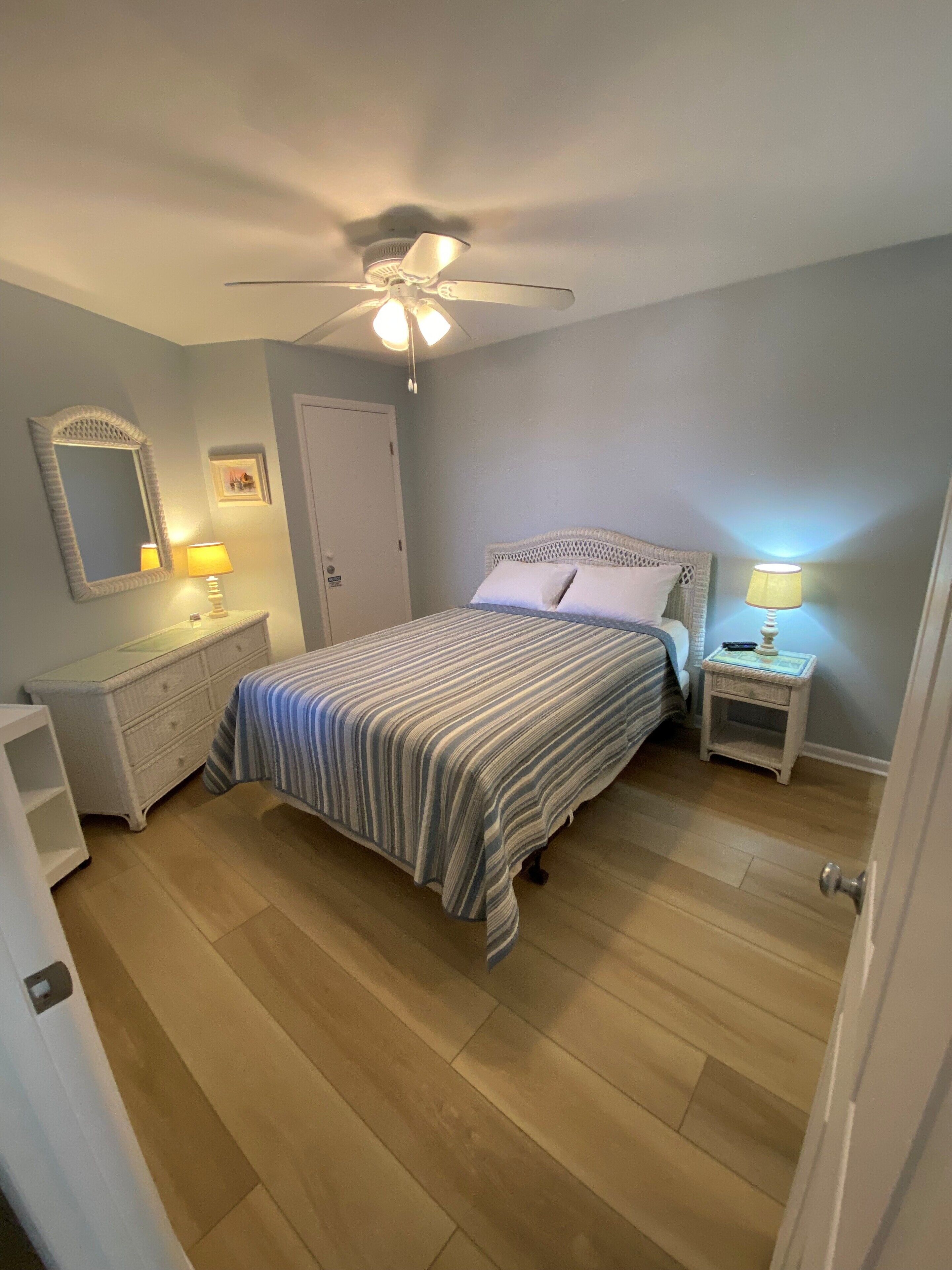 A bedroom with a bed, two bedside tables, a ceiling fan, a mirror, and a door.