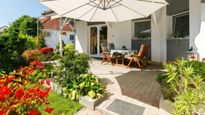 Une terrasse avec une table et des chaises sous un grand parasol, entourée de plantes en pot et d’un jardin.