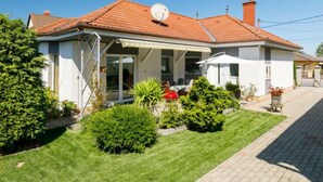 Une maison à deux étages, avec un toit recouvert de tuiles rouges, un porche abrité et un petit jardin agrémenté de diverses plantes et d’un banc.