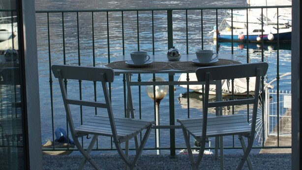 Un balcón con mesa y sillas con vista a un lago con barcos, un pueblo al fondo y montañas.