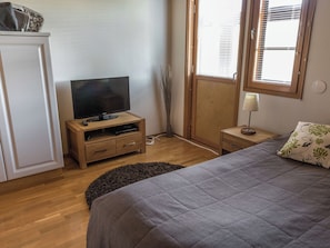 Un dormitorio con cama, televisor sobre un mueble de madera, mesita de noche con lámpara y ventana con persianas.
