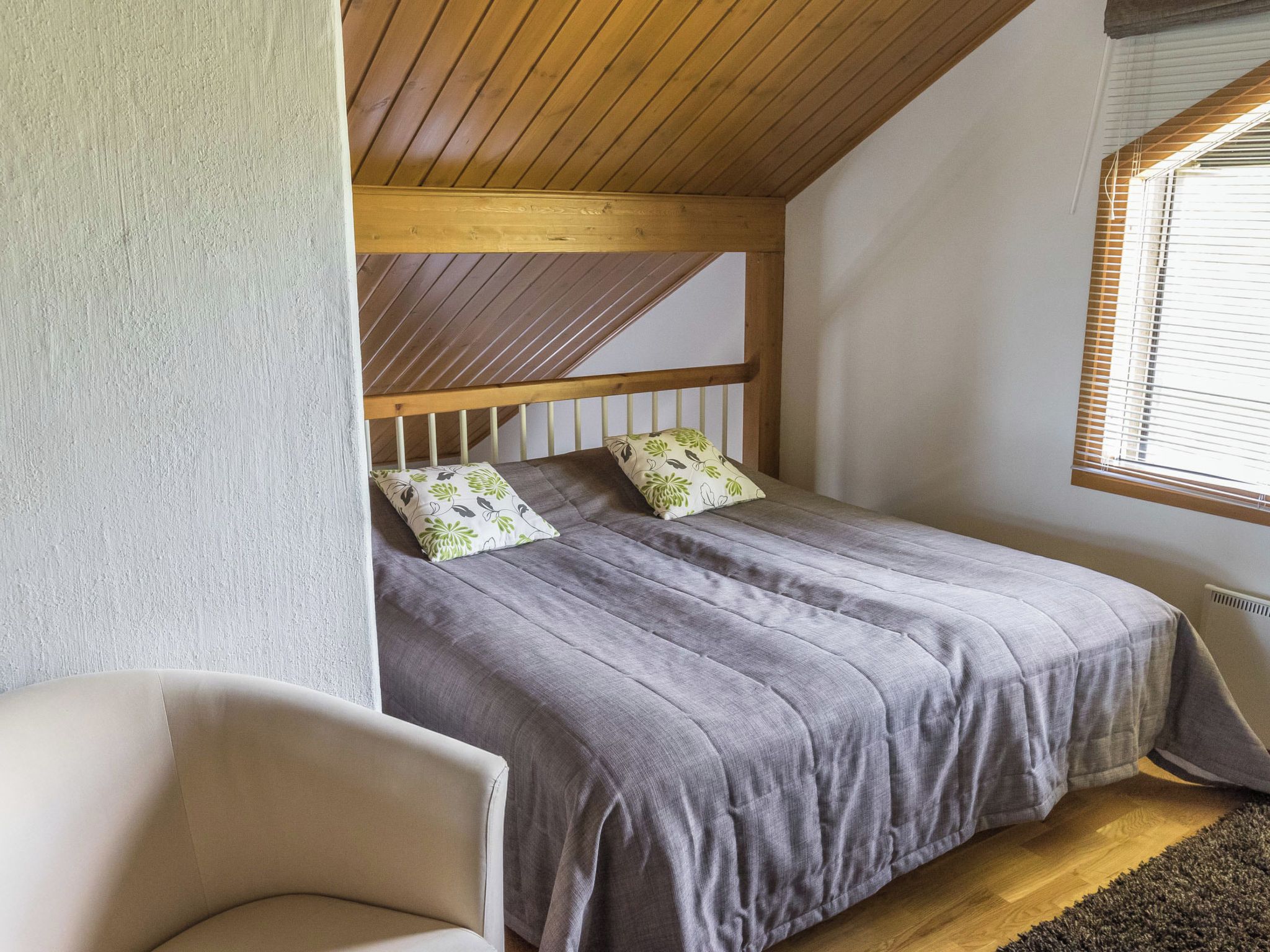 Ein Schlafzimmer mit geneigter Decke, einem Bett mit grauer Bettwäsche, zwei Kissen mit Blumenmuster, einem Holz-Kopfende, einem Stuhl und einem Fenster mit Jalousien.